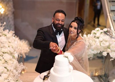 Two people dressed in formal wedding attire are cutting a three-tiered white cake surrounded by white flowers. They are smiling and standing in front of a staircase at The Cable Center, an elegant Denver event venue.