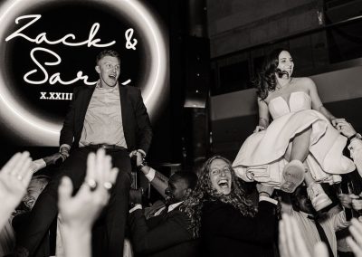 A black and white image of a couple being lifted on chairs by guests at a wedding celebration at The Cable Center, an elegant Denver event venue. A neon sign in the background reads “Zack & Sara X.XXIII.”