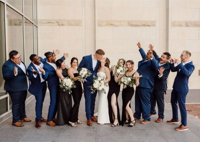 A wedding party of twelve, dressed in formal attire and standing in a row, celebrates as the bride and groom kiss in the center. The men wear blue suits, and the women wear black dresses. This beautiful moment takes place at The Cable Center, an elegant event venue in Denver that adds a perfect touch of sophistication to their special day.