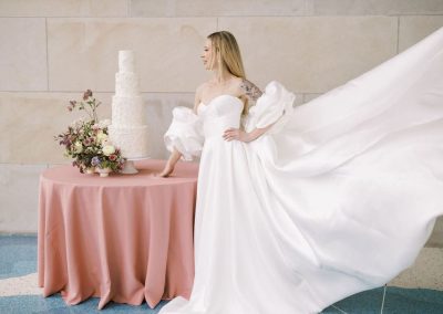A bride in a strapless white gown with billowing sleeves stands beside a four-tiered wedding cake on a round table, covered with a pink tablecloth and floral arrangement. The train of her wedding gown flows high up behind her.