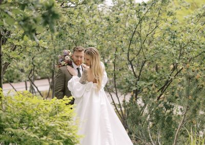 A bride in an elegant white gown and groom in a tailored suit share a moment surrounded by lush green trees and bushes at The Cable Center event venue in Denver.