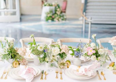 A table is elegantly set at The Cable Center, a unique Denver event venue, featuring white plates, gold cutlery, and floral centerpieces with white, pink, and purple flowers. Folded pink napkins are placed on each plate.