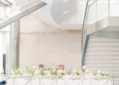 A long, elegant dining table with white chairs is set with floral arrangements in the modern, bright, and spacious room featuring a sweeping staircase and minimalist decor at The Cable Center, perfect for an elegant Denver event.