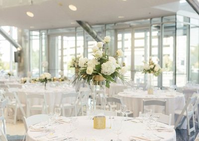 An elegant banquet hall with round tables covered in white tablecloths, each adorned with floral centerpieces and table settings, illuminated by natural light from large windows, providing a unique charm at The Cable Center event venue in Denver.