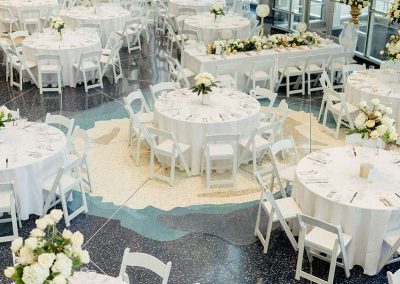 The spacious Denver event venue The Cable Center is set up for an elegant gathering with round tables covered in white tablecloths, white chairs, and floral centerpieces. Natural light streams in from large windows.