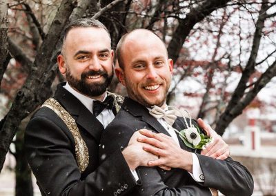 Two smiling men in elegant formal wear pose together at their outdoor wedding. One man embraces the other from behind, both wearing suits and boutonnières. Their joy and style emanate effortlessly. There are leafless trees in the background.