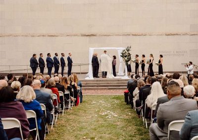 Elegant outdoor wedding ceremony with guests seated on either side of a grassy aisle, facing a couple and officiant under a white canopy, flanked by their bridesmaids and groomsmen at The Cable Center event venue in Denver.