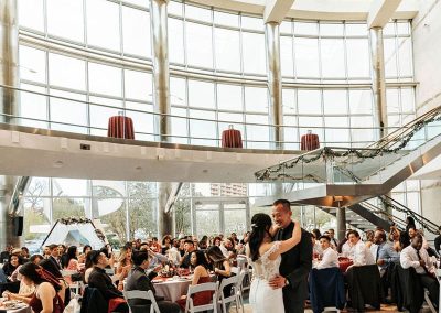 A recently-married couple dances in the center of The Cable Center’s large Daniels Great Hall, with floor-to-ceiling windows. Wedding guests are seated at tables around the room, watching the couple’s first dance. The elegant setting provides a unique backdrop for their enchanting moment.