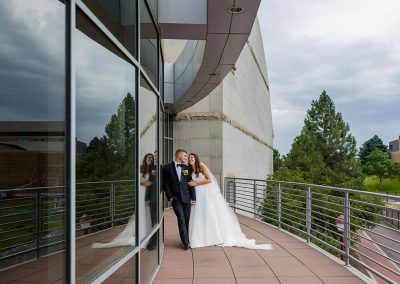 A couple in wedding attire stands closely together on an outdoor balcony at The Cable Center in Denver. There is a modern building and trees in the background under cloudy skies. Their reflection is visible in the glass wall beside them, adding an elegant touch to the unique event venue.