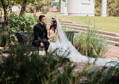 A bride and groom sit on a park bench, surrounded by greenery, with the bride's veil trailing beside them, creating an elegant scene at The Cable Center, a unique Denver event venue.