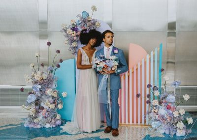 A young couple stands together at the elegant, floral-decorated Cable Center event venue in Denver. There are pastel geometric design elements in the background. The bride wears a white dress, and the groom is in a blue suit holding a bouquet.