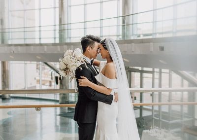 A couple dressed in wedding attire embraces in the modern, spacious Cable Center event venue with large windows. The bride holds a bouquet and wears a long veil and gown, while the groom wears a dark suit.