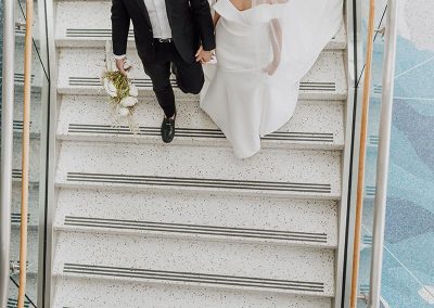 A couple dressed in wedding attire descends a staircase, holding hands. The bride wears an elegant white gown and veil, while the groom is in a sleek black suit. Their moment is captured beautifully at The Cable Center, a unique Denver event venue.