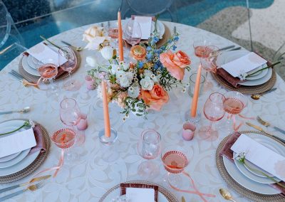 An elegant round table set for six with a white tablecloth, pink glassware, and orange taper candles graces the unique decor of The Cable Center event venue in Denver. The centerpiece boasts various flowers, including roses, while place settings feature menus and pink ribbon accents.