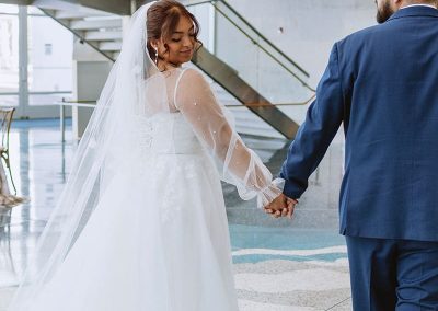 A bride in a unique, white gown and veil holds hands with a groom in an elegant blue suit as they walk together in The Cable Center’s Daniels Great Hall, a modern Denver event venue with glass railings and stairs.