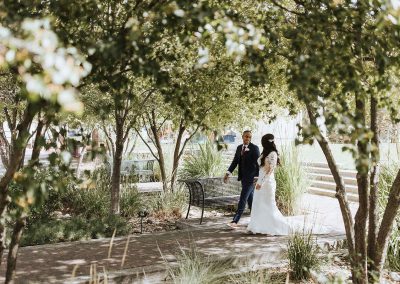 A couple in wedding attire walks hand-in-hand through a shaded park pathway, surrounded by trees and greenery, setting the scene at the perfectly elegant Cable Center event venue in Denver, CO.