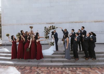 An elegant wedding party poses on steps of The Edward S. Rogers Amphitheater at The Cable Center, a unique Denver event venue. The bride and groom stand in the center with bridesmaids in red dresses and groomsmen in grey suits cheerfully raising their arms.