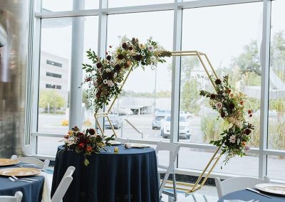 A wedding reception setup at The Cable Center, an elegant Denver event venue with large windows. There is a hexagonal floral arch, round tables with navy blue tablecloths, and white chairs.