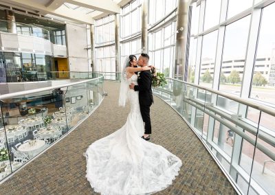 In The Cable Center, a Denver event venue, a bride and groom share a moment on a curved balcony in an atrium with large windows. The bride wears a long, elegant white dress, and the groom is in a black suit. Unique tables are set up below them.