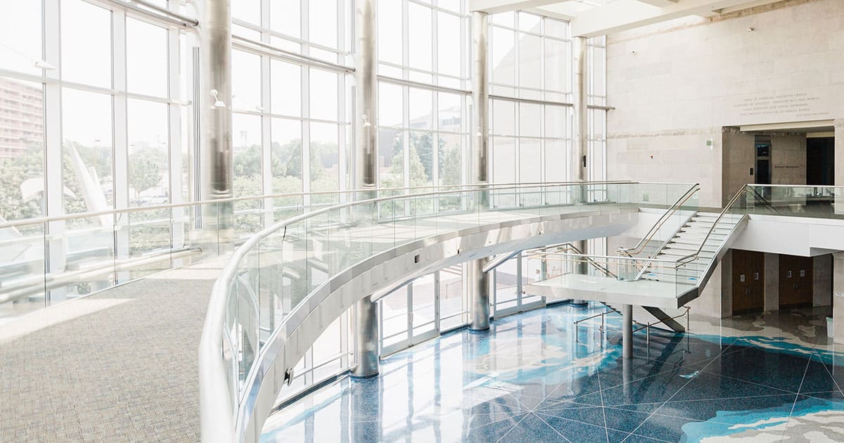 The Cable Center in Denver features a spacious modern atrium with large glass windows, a curved glass-railed walkway, and a staircase descending to a beautifully patterned blue floor.