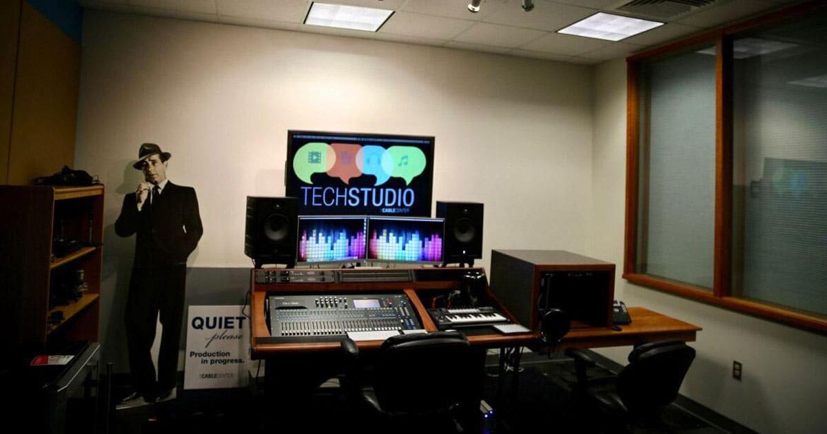 A large mixing console, computer monitors displaying graphics, and a vintage-style figure cutout. A sign reads "Quiet please, recording in progress," creating an atmosphere of creativity and precision at The Tech Studio at The Cable Center in Denver.